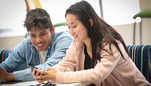 Two students sitting at a table, smiling and looking at a phone